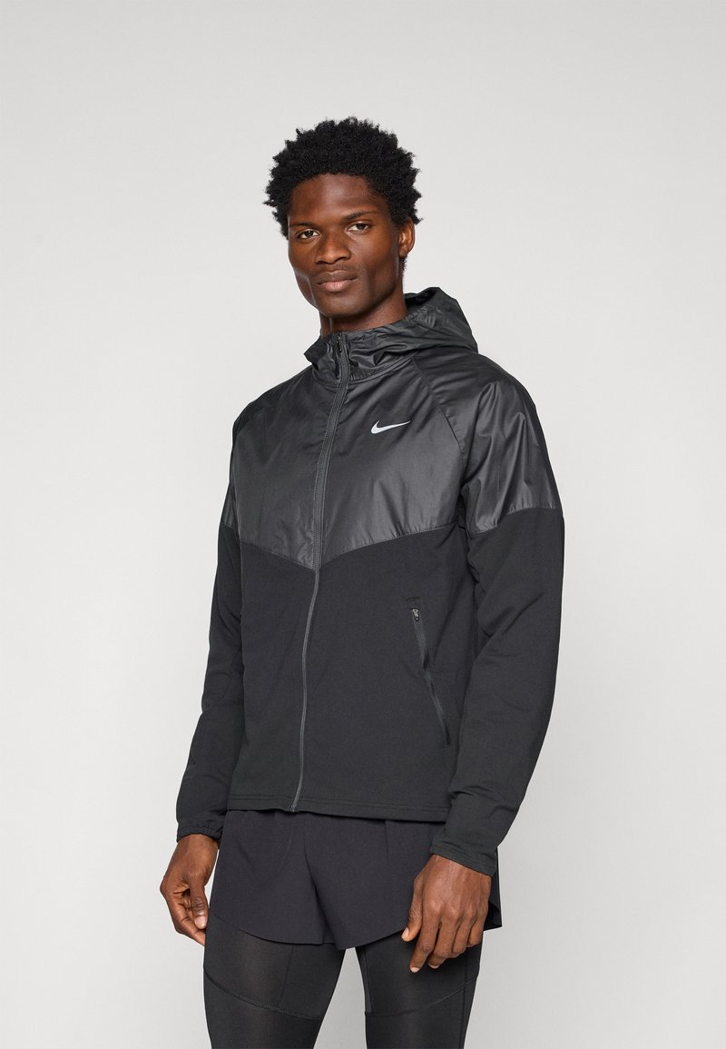 Czarna sportowa kurtka z błyszczącą czarną górną częścią, kapturem, zapięciem na zamek, bocznymi kieszeniami oraz logo Nike na piersi.