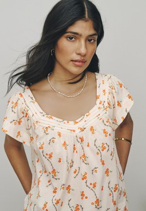 Młoda kobieta z długimi czarnymi włosami ubrana w kremową kwiecistą bluzkę, perłowy naszyjnik, złote kolczyki-koła oraz złotą bransoletkę, pozująca na jednolitym tle.