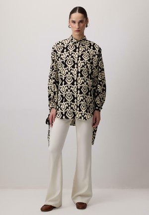 Black and beige patterned oversized shirt with a high collar and long sleeves, paired with cream wide-leg pants and brown shoes.
