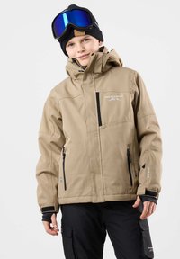 Beige Skijacke mit Reißverschluss, Taschen und verstellbaren Bündchen. Getragen über schwarzen Cargo-Hosen, kombiniert mit schwarzer Mütze und Schutzbrille.