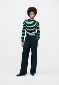 Ριγέ navy και πράσινο πουλόβερ με μακριά μανίκια, σε συνδυασμό με φαρδιά σκούρα παντελόνια. Λεπτή καφέ ζώνη και μαύρα παπούτσια ολοκληρώνουν την εμφάνιση.