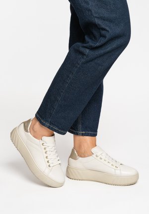 Weiße Plateausneaker mit goldfarbener Absatzverzierung, getragen zu dunkelblauen, aufgekrempelten Jeans vor einfachem Hintergrund.