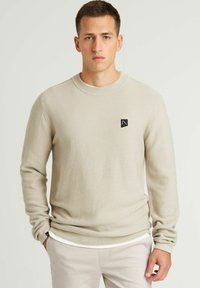 Maglione lavorato a maglia beige con scollatura rotonda e patch logo nero sul lato sinistro del petto. Maniche lunghe e polsini a coste. Vestibilità sartoriale.