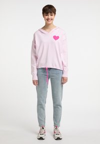 Lichtroze hoodie met een roze hartontwerp, trekkoord in de taille en geribde manchetten. Gemonstreerd met lichtblauwe jeans en gemusterde sneakers.