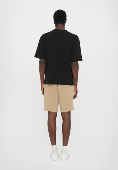 Tricou negru, supradimensionat, din bumbac, purtat cu pantaloni scurți bej. Adidași și șosete albe completează ținuta. Design simplu, casual.