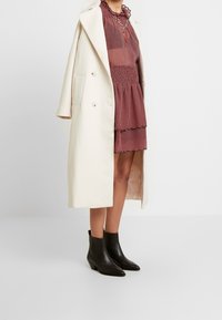 Lång krämfärgad kappa, åtsittande röd klänning med prickar och volang vid halsen, samt svarta ankelskor med spetsiga tår och en lätt klack.