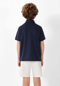 Camisa polo de color azul marino con mangas cortas y cuello de canalé, combinada con pantalones cortos de color claro. Presenta una pequeña etiqueta lateral.
