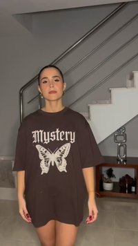 Sötét haját kontyba tűzött nő fekete, túlméretezett „Mystery” feliratú és pillangós mintás pólót visel, beltéren, lépcső és polc közelében áll.