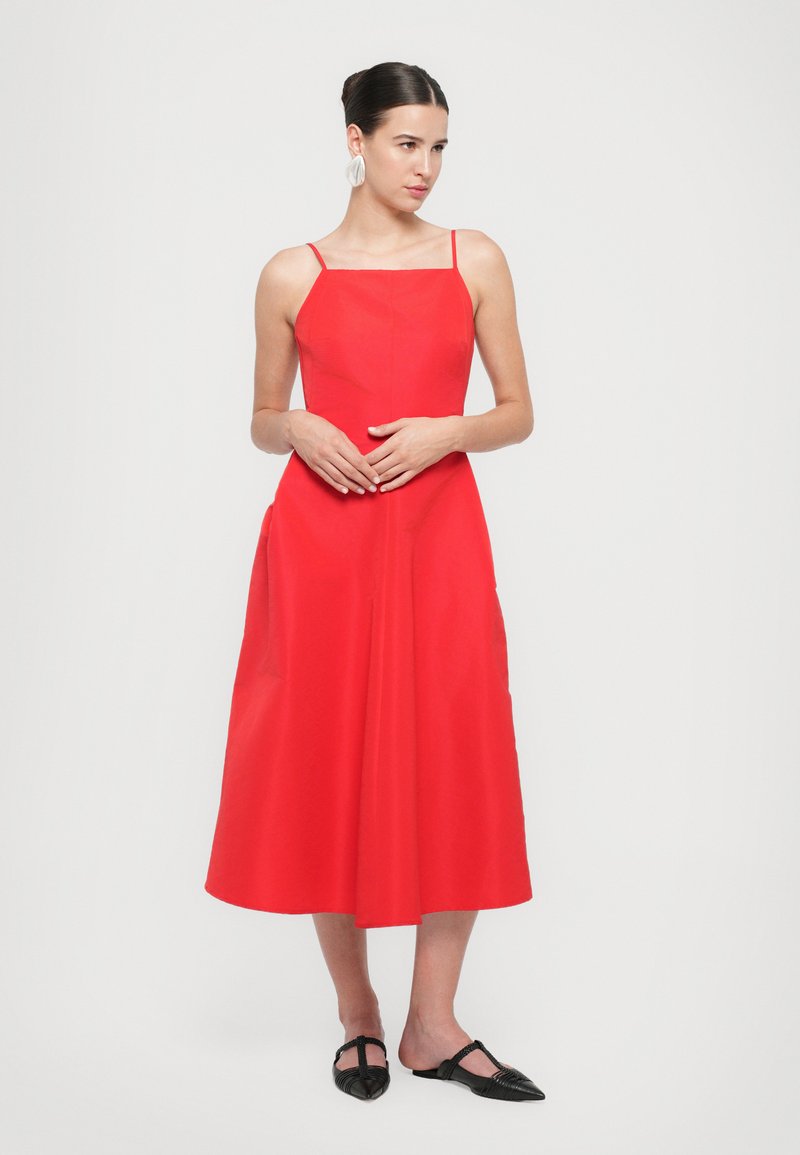 Modèle dans une robe sans manches rouge vif se tient devant un fond uni, portant des boucles d'oreilles élégantes et des ballerines noires, avec une expression pensive.