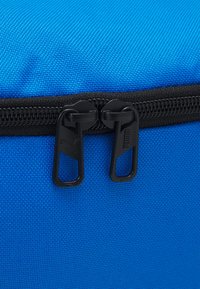 Sac en textile bleu avec une fermeture éclair noire robuste à double tirette et une texture de surface lisse. Les accents comprennent un design de tirette de marque.