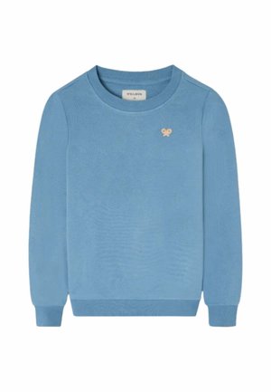 Niño con sudadera azul, pantalones cargo beige, deportivas azul marino y una gorra de béisbol amarilla y gris, de pie y mirando hacia un lado.