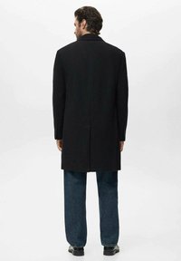 Abrigo negro de corte ajustado, con una abertura simple en la parte posterior y solapas con muesca, combinado con jeans rectos azules y zapatos negros.