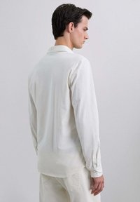 Camisa blanca de manga larga con cuello, confeccionada en una tela suave. Presenta un corte holgado y puños abotonados, con una textura lisa y sin adornos.