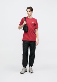 Mladi moški, oblečen v rdečo majico Nike, črne hlače, sive superge Nike in z ruto črno torbico čez eno ramo.