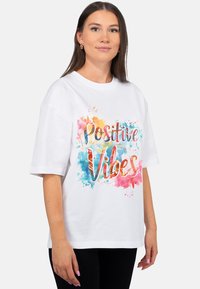 Witte katoenen T-shirt met korte mouwen, bedrukt met de tekst "Positive Vibes" in een kleurrijke aquarelstijl en spatten in roze, blauw en oranje.