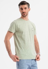 Camiseta de algodón verde claro con manchas negras, cuello redondo, mangas cortas y un pequeño diseño de logo en el pecho.