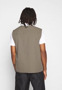 Olivgrön ärmlös väst i ett slätt material, med hög krage och en liten ögla på baksidan. Bärs över en vit skjorta.