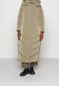 Abrigo acolchado largo en beige claro, con un cuello alto, capucha y costuras en forma de chevron. Combinado con botas oscuras hasta el tobillo y pantalones marrones holgados.