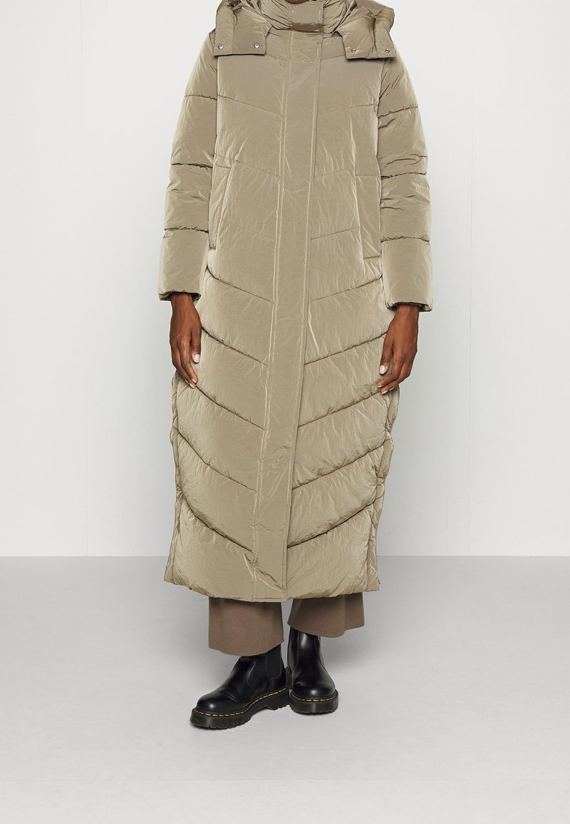 Abrigo acolchado largo en beige claro, con un cuello alto, capucha y costuras en forma de chevron. Combinado con botas oscuras hasta el tobillo y pantalones marrones holgados.