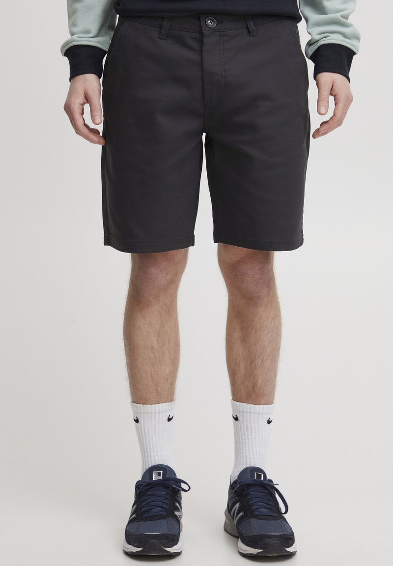 Sorte shorts laget av slitesterkt stoff, med knappelukking, sidelommer, og rett snitt, brukt med hvite ankelsokker og sneakers.