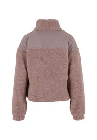 Jakna s cropped krojem, obložena flisom, u mekoj bež boji, s glatkom kontrastnom tkaninom na ramenima. Ima visoki ovratnik i elastične manšete.