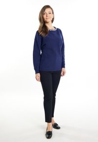 Marineblauer Strickpullover mit langen Ärmeln und rundem Ausschnitt, verziert mit floralem Akzent. Kombiniert mit schwarzen Slim-Fit-Hosen und schwarzen Slippern.