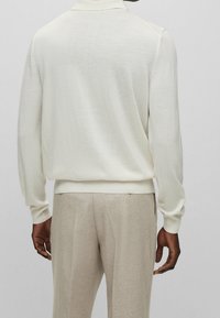 Pull en tricot crème clair avec col, manches longues et ourlet côtelé, associé à un pantalon beige, mettant en valeur une texture lisse.
