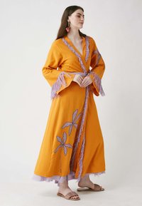 Largo vestido envolvente naranja con mangas acampanadas, acentos de flecos morados y bordados florales. Capas con un forro lavanda en el dobladillo.