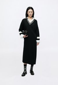 Črna prevelika obleka v stilu puloverja z belim ovratnikom, črtastimi manšetami in kontrastnim V-izrezom. Kombinirana s črnimi loaferji in črtastimi nogavicami.