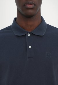 Gros plan d'une personne à la peau foncée portant un polo bleu marine foncé avec une patte à deux boutons et un texte brodé subtil sur la poitrine.