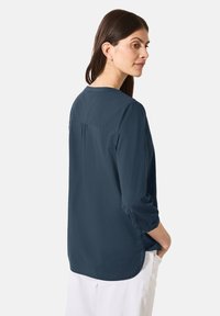 Blusa a maniche lunghe blu navy con scollatura rotonda, polsini con bottone e dettaglio di piega sul retro. Tessuto morbido, vestibilità rilassata.
