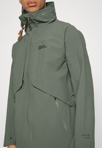 Homme portant une veste verte Jack Wolfskin avec un col haut et plusieurs poches zippées, vue partielle de la poitrine à la tête.