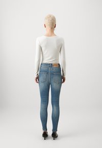 Jeans azul de tiro alto y ajustados con un lavado desgastado, cinco bolsillos y una etiqueta de cuero marrón en la parte posterior. Combinados con una blusa blanca de manga larga.
