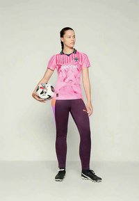 Mulher em uniforme de futebol rosa e roxo segurando uma bola de futebol, de pé contra um fundo claro e liso, olhando para o lado.