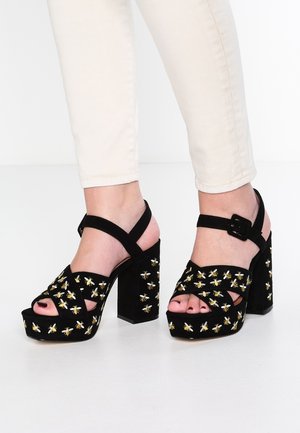 Zwarte plateauhakken met grove hakken, enkelbandjes en gele bijenborduursels, gedragen met lichte broek op een witte vloer.
