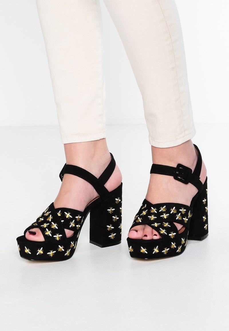 Chaussures à plateforme noires à talons épais, avec brides à la cheville et broderie d'abeille jaune, portées avec un pantalon clair sur un sol blanc.