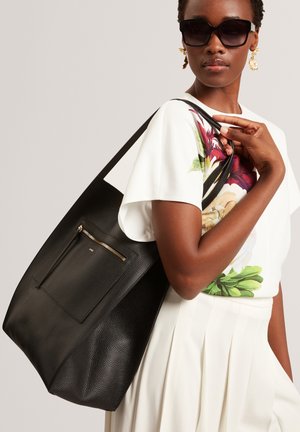 Zwarte leren tote bag met een gewelfde textuur, voorzien van een ritszak en schouderband. Het model draagt een top met bloemenpatroon en een zonnebril.