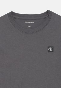 Tricou din bumbac gri cu guler rotund, având un mic logo alb-negru "cK" pe partea stângă a pieptului. Textură netedă, cu cusături minime.