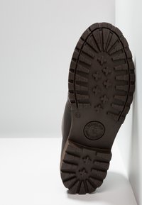 Pruun kummist saapasseene, sügavate mustritega, ümmargune konts, ringikujuline logo ja suuruse märgistus; tekstureeritud haardumise jaoks, näidates tugevat disaini.