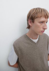 Light brown, sleeveless knit vest with a V-neck design worn over a white T-shirt. Smooth texture against a plain wall backdrop.