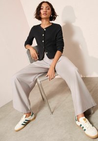 Crni rebrasti kardigan s okruglim izrezom i kopčanjem, uparen s svijetlosivim širokim hlačama i bijelim tenisicama s zelenim naglascima.