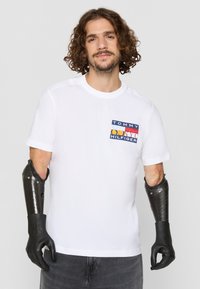 Hvid kortærmet t-shirt med et multicolor firkantet logo-patch med teksten "TOMMY HILFIGER." Modellen har en sort protesearm.