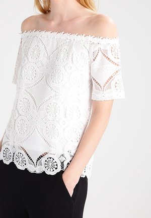Kvinde iført en hvid off-shoulder blondebluse med blomsterprint og halværmer, kombineret med sorte bukser, hånd i lommen.