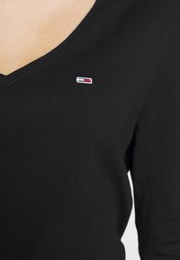 Svart V-ringad topp i mjuk bomull, med en liten broderad logotyp i röd, vit och blå på vänster bröstområde.