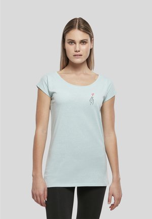 Jeune femme aux cheveux raides porte un t-shirt bleu clair avec une petite main brodée formant un symbole de cœur sur la poitrine.