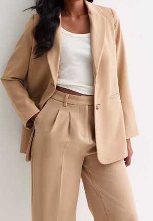 Vrouw die een beige op maat gemaakte blazer draagt en bijpassende hooggetailleerde geplooide broek met wit geribd cropped top op een effen achtergrond.