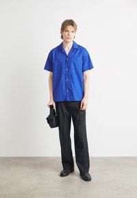 Camisa azul de manga curta com botões e um bolso, combinada com calças largas pretas e sapatos pretos, segurando uma pequena bolsa preta.