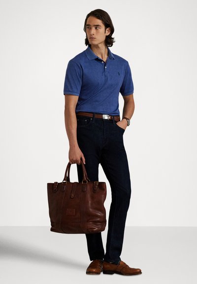 Muž v modrom polokošeli a tmavých džínsoch drží veľkú hnedú koženú kabelku, na nohe má hnedé mokasíny a na ruke hodinky, stojí pred jednoduchým pozadím.