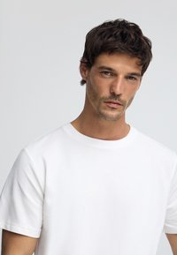 Camiseta de algodón blanca con cuello redondo y mangas cortas. Textura suave, diseño sencillo, sin patrones ni acentos visibles.