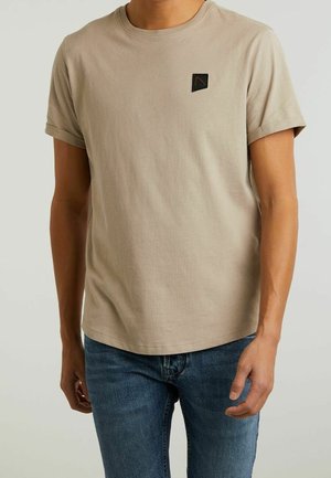 Mężczyzna ubrany w beżowy t-shirt z krótkim rękawem z małym czarnym logo na piersi, w połączeniu z niebieskimi dżinsami, stojący na jednolitym tle.